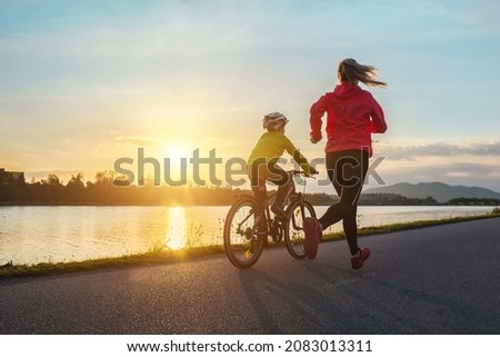 Happy mother and son go in sports outdoors. Boy rides bike in helmets, mom runs on sunny day. Silhouette family at sunset. Fresh air. Health care, authenticity, sense of balance and calmness Royalty-Free Stock Photo #2083013311