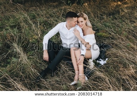 Stylish groom and cute blonde bride with curly hair in a white dress are sitting on a rug on the yellow grass in nature and hugging. Wedding portrait of beautiful newlyweds.
