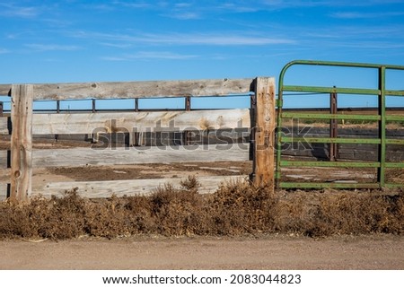 An old wooden fence connected to a newer green pipe gate.  Blue sky with wispy cloud in the sky.  Tan dirt with brown weeds at the base create a rustic scene. 