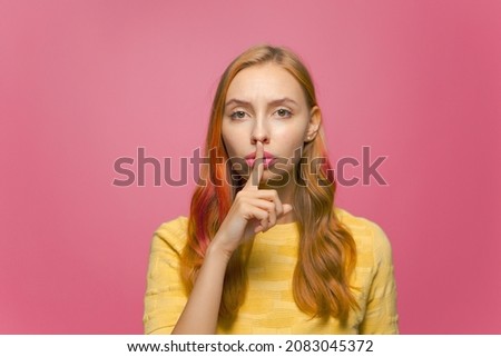 Beautiful serious young girl ask to be quiet and keep secret, showing silence gesture on pink studio background