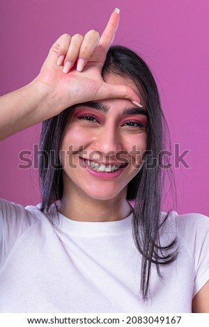 Arabic female model showing loser sign on her forehead. Isolated on pink background.