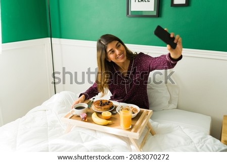 Beautiful woman taking a selfie with her smartphone while eating breakfast in bed during a lazy morning