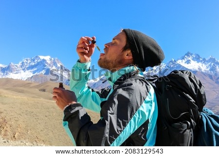Young caucasian male with a beard wearing a windbreaker and a black beanie taking CBD oil from a dripper high up in the Himalaya mountains. Sports enhancement supplement for trekking in high altitude. Royalty-Free Stock Photo #2083129543