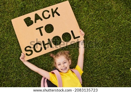 From above of smiling little girl with backpack lying on green grassy lawn holding carton poster saying Back to school
