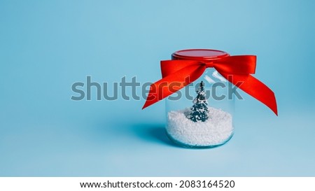 Christmas tree in snow closed in transparent jar with red bow tie satin ribbon on blue background. Creative Xmas eve or Happy New Year celebration concept. Minimal winter holidays greeting card.