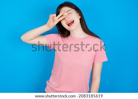 Caucasian woman wearing pink T-shirt over blue background Doing peace symbol with fingers over face, smiling cheerful showing victory