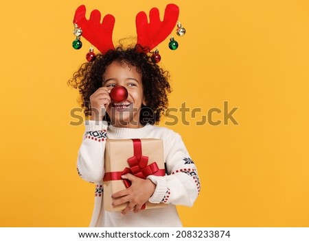 Cheerful little african american boy with wrapped gift holding red christmas tree ball in front of his nose, wearing warm   xmas sweater and deer horns on head, posing against yellow studio background