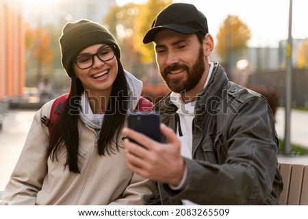 Young happy caucasian lover woman and man embracing together tenderly while sitting at the bench at the street and making selfie with each other. Love and date concept 