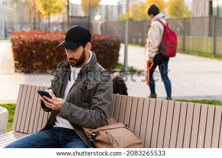 Mirthful man sitting at the bench and smiling while holding his phone and looking at the screen with pleasure smile. Human and technologies concept