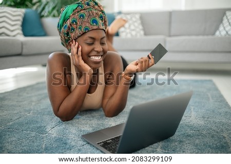 Black African woman wearing traditional headscarf, making online shopping purchase using laptop and credit card. Laying on rug in lounge at home
