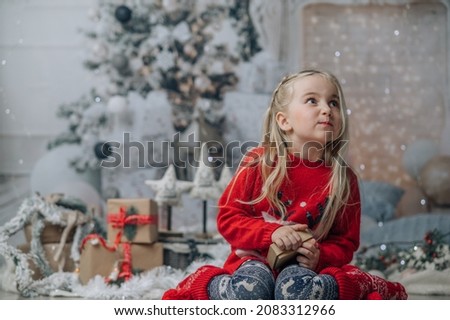 Little girl opening christmas present, looking at gift box with smile hild wearing Chrismas jumper posing on floor near fireplace and xmas tree.