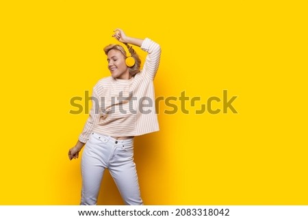 Joyful caucasian woman raises hand in dance move while listening music in headphones. Isolated over yellow bckground with copy space