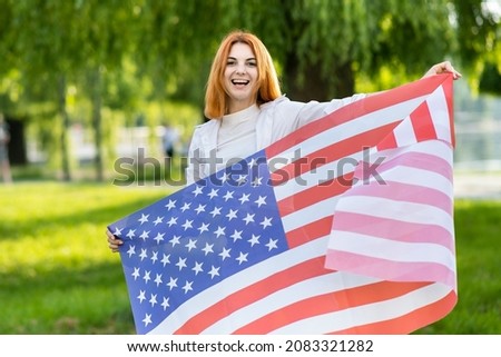 Young red haired woman holding USA national flag standing outdoors in summer park. Positive girl celebrating United States independence day. International day of democracy concept.