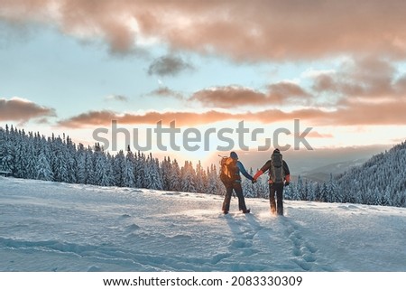 Romantic couple of tourists dressed in warm winter sportswear with tourist backpacks walking with trekking poles in the snowy pine mountains in an incredible sunset. Family, rest. Royalty-Free Stock Photo #2083330309