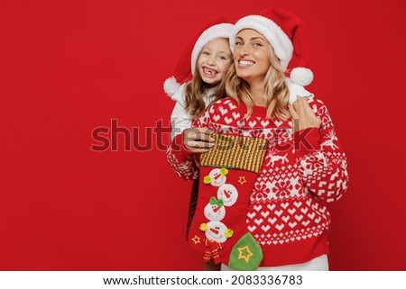 Young happy woman in sweater hat have fun with child baby girl 6-7 years old. Mommy little kid daughter look at gift sock stocking isolated on plain red background studio New Year love family concept