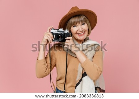 Traveler tourist smiling mature elderly senior lady woman 55 years old wears brown shirt hat scarf hold retro vintage camera take photo isolated on plain pastel light pink background studio portrait