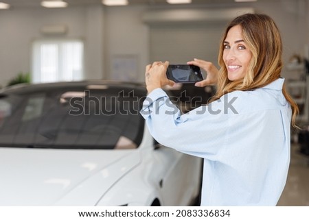 Happy woman customer female buyer client wears blue shirt do photo on mobile phone choose auto want buy new automobile in car showroom vehicle salon dealership store motor show indoor. Sales concept