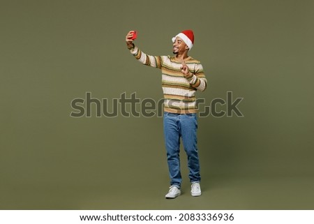 Full body young african man in sweater red Christmas Santa Claus hat doing selfie shot on mobile cell phone show v-sign isolated on plain green khaki background Happy New Year 2022 celebration concept