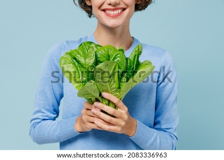 Young cropped close up smiling surprised caucasian woman 20s in casual sweater hold green lettuce leaves isolated on plain pastel light blue background studio portrait. People lifestyle food concept