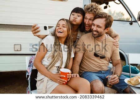 Multiracial two couples laughing and taking selfie photo while leaning on trailer outdoors
