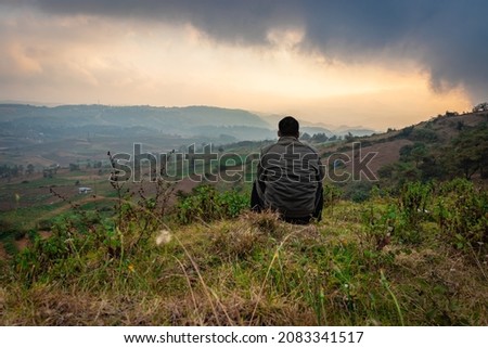 young man sitting on mountain top with misty hill range background and dramatic sky at morning image is taken at badda peak shillong meghalaya india.