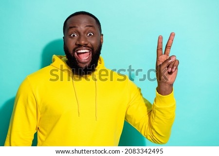 Photo of brunette hooray young beard guy show v-sign wear yellow hoodie isolated on teal color background