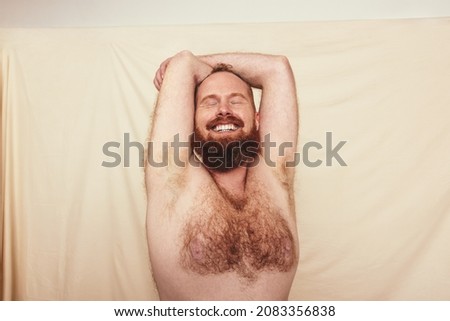 Embracing everything natural. Excited young man smiling with his eyes closed while standing shirtless against a studio background. Self-loving man embracing his natural and hairy body.