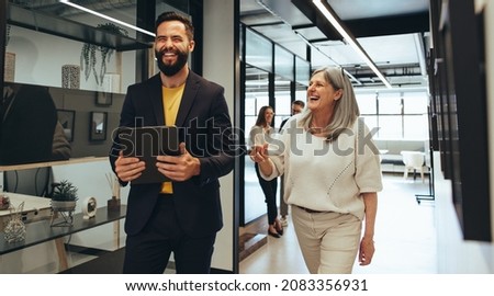 Laughing entrepreneurs walking together in an office. Two happy business colleagues sharing a laugh while having a discussion. Diverse businesspeople working together on a project. Royalty-Free Stock Photo #2083356931