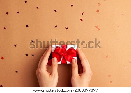 Human hands puts a white paper gift with a red satin ribbon bow on brown background with red stars shapes. Christmas Holidays concept flat lay with copy space.