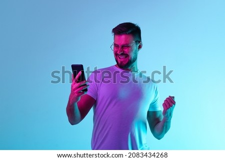 Pleasant news. Portrait of happy excited man joyfully looking at phone screen isolated over blue background in neon lights. Concept of human emotions, facial expression, lifestyle. Copy space for ad