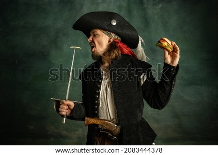 Portrait of one brutal man, medieval pirate in hat pricking fries and holding burger isolated over dark background. Combination of medeival and modern styles. Concept of history. Copyspace for ad.