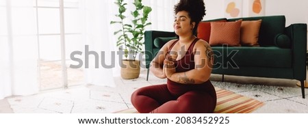 Healthy female in practicing meditation yoga at home. Body positive woman exercising with eyes closed and hands joined while sitting cross legged in living room. Royalty-Free Stock Photo #2083452925