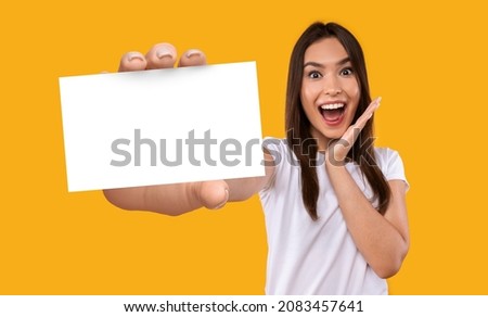 Portrait of excited young Caucasian woman holding blank white paper card with mockup for your business information, orange studio background. Banner design. Template for company data