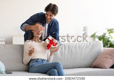 Smiling young european husband closes eyes to his wife and gives gift box in living room interior. Birthday surprise, valentine day and anniversary, romantic date at home during covid-19 quarantine