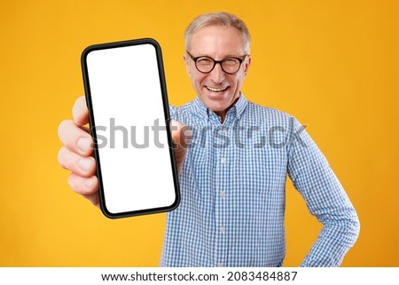 App Advertisement. Excited Adult Man In Glasses Showing Big White Empty Smartphone Screen Close To Camera Recommending Website Posing At Yellow Orange Studio. Check This Cellphone Display Mock Up