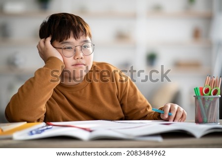 Asian schooler chubby boy dreaming about something while doing homework, sitting at desk with notebooks and stationery, taking notes, touching his head, looking at copy space, home interior