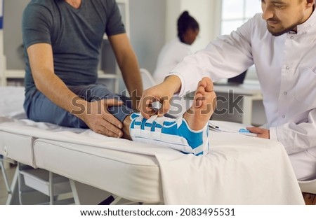 Traumatologist fixes orthopedic bandage on leg of male patient with fracture of ankle joint. Close up on examination couch injured leg of patient receiving first aid in trauma department. Royalty-Free Stock Photo #2083495531