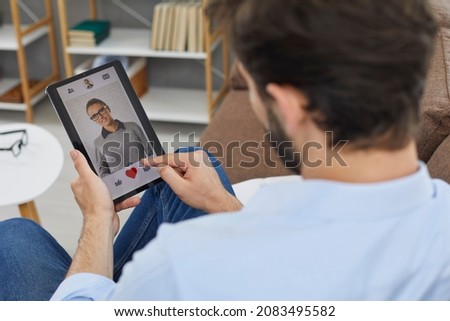 Man on digital tablet looks at profile photo of attractive young woman in dating app and presses red heart like button. Concept of finding love online. Close-up, selective focus.