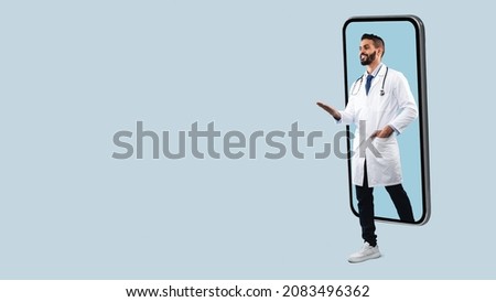 Telemedicine. Young Happy Male Middle Eastern Doctor In White Uniform With Stethoscope Coming Out Big Cell Phone Screen, Stepping Walking Off Gadget Monitor, Showing Free Copy Space, Panorama Banner Royalty-Free Stock Photo #2083496362