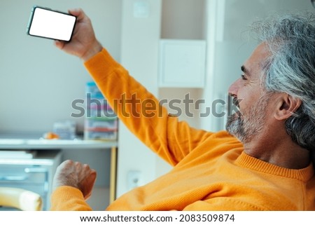 Smiling Mature Man Taking Selfie on a Mobile Phone While Sitting On a Dentist's Chair. 
Male patient in orange sweater holding his smartphone with blank screen while waiting to be seen by the dentist.