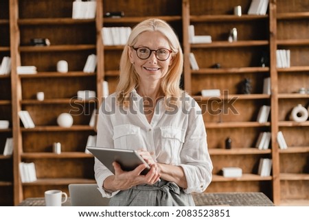 Smart mature middle-aged businesswoman freelancer tutor teacher boss ceo holding digital tablet looking at the camera while working remotely in home office