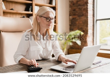 Middle-aged mature businesswoman boss financial advisor counting on calculator funds, money, paying domestic bills, debts, credit loan using laptop in office Royalty-Free Stock Photo #2083529695