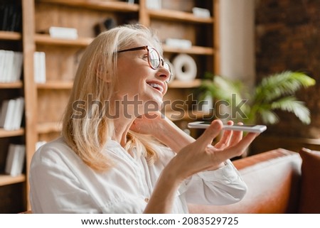 Mature caucasian businesswoman freelancer using cellphone recording voice message online with business partners, using voice assistant at home office remotely.