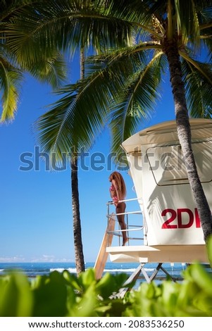 Charming curly African-American dark-skinned young woman, professional surfer stay on lifeguard tower with surfboard with palm trees and ocean at the background