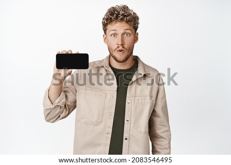 Image of amazed guy showing mobile phone screen horizontally, staring impressed at camera while demonstrating something cool on smartphone app, white background