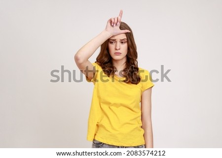 Loser! Portrait of displeased upset woman of young age in yellow T-shirt showing loser gesture, L finger symbol on head, worried about failure, mistake. Indoor studio shot isolated on gray background.