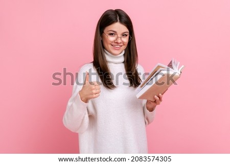 Beautiful satisfied woman showing thumbs up gesture holding and reading book, likes genre and plot, wearing white casual style sweater. Indoor studio shot isolated on pink background.