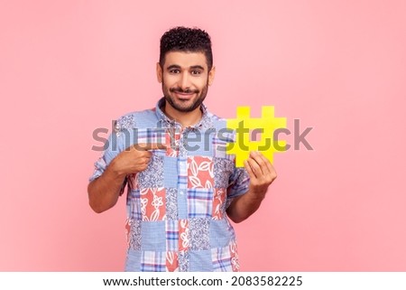 This is internet trend. Portrait of bearded man with dark hair in blue casual shirt smiling and pointing at big hashtag sign, sharing viral content. Indoor studio shot isolated on pink background.