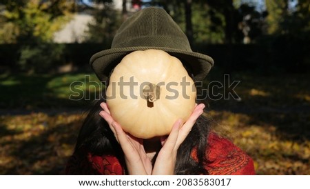Girl with a hat on her head holding a round pumpkin in front of her face. Funny autumn picture with a pumpkin.
