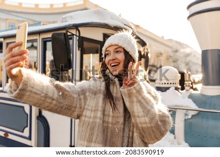 Cheerful fair-skinned young girl makes selfie on phone showing two fingers to camera. Brunette with toothy smile, wearing hat and coat. Holidays, season and leisure concept.